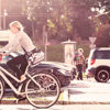 A female cyclist rides through town with cars, a bus and pedestrians in the background.